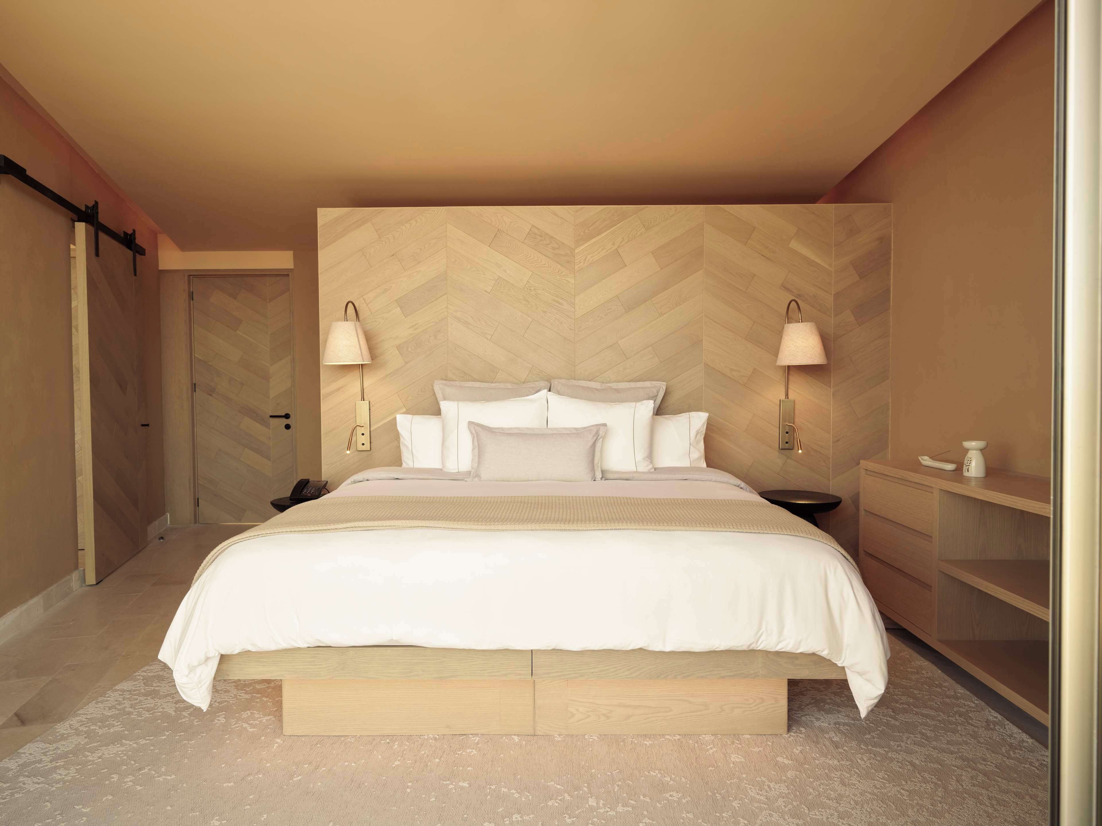 Un dormitorio moderno con cabecera de diseño en patrón espiga, una cama grande con ropa de cama blanca, dos lámparas de noche y una mesita de noche con lámpara.