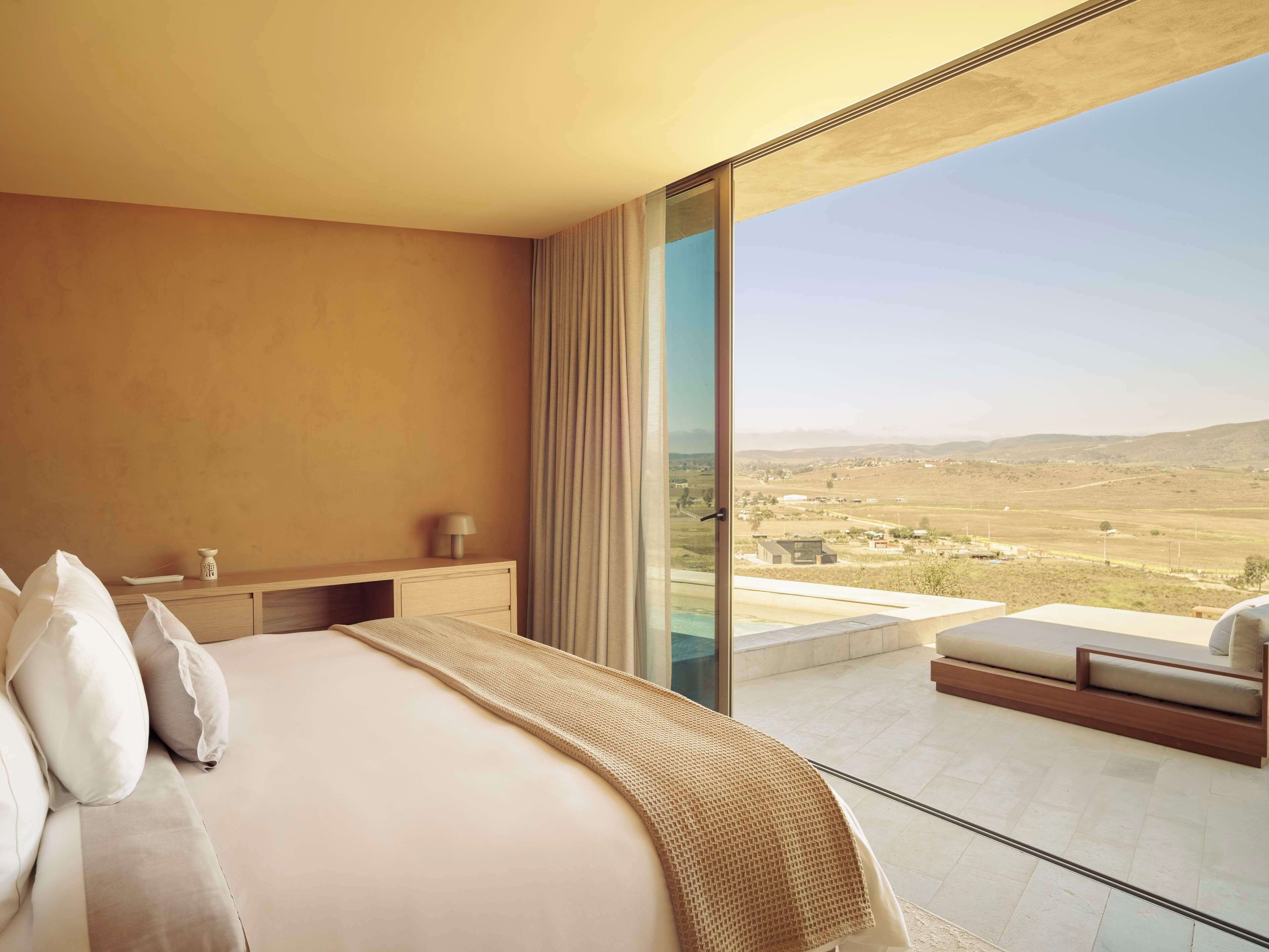 A bedroom with a large bed, a bedside table, a lamp, and a view of a landscape through a glass door.
