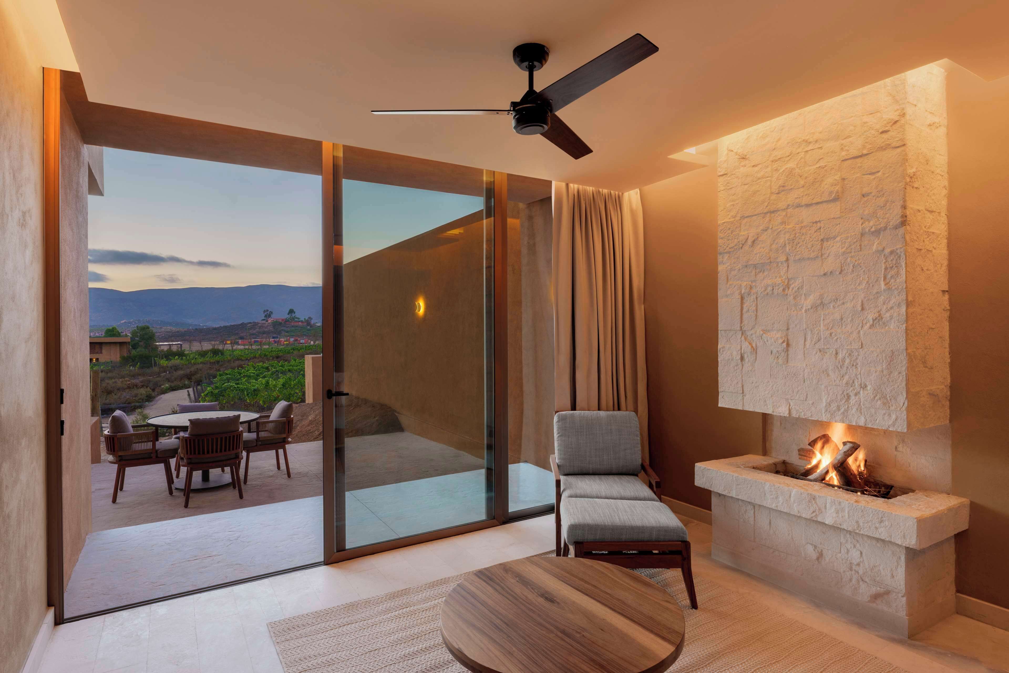 Una sala de estar moderna con chimenea, ventilador de techo y vistas a un jardín ajardinado a través de amplios ventanales.
