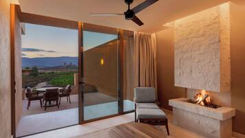 Una sala de estar moderna con chimenea, ventilador de techo y vistas a un jardín ajardinado a través de amplios ventanales.