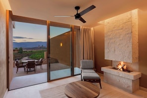 A modern living room with a fireplace, a ceiling fan, and a view of a landscaped area through large glass doors.