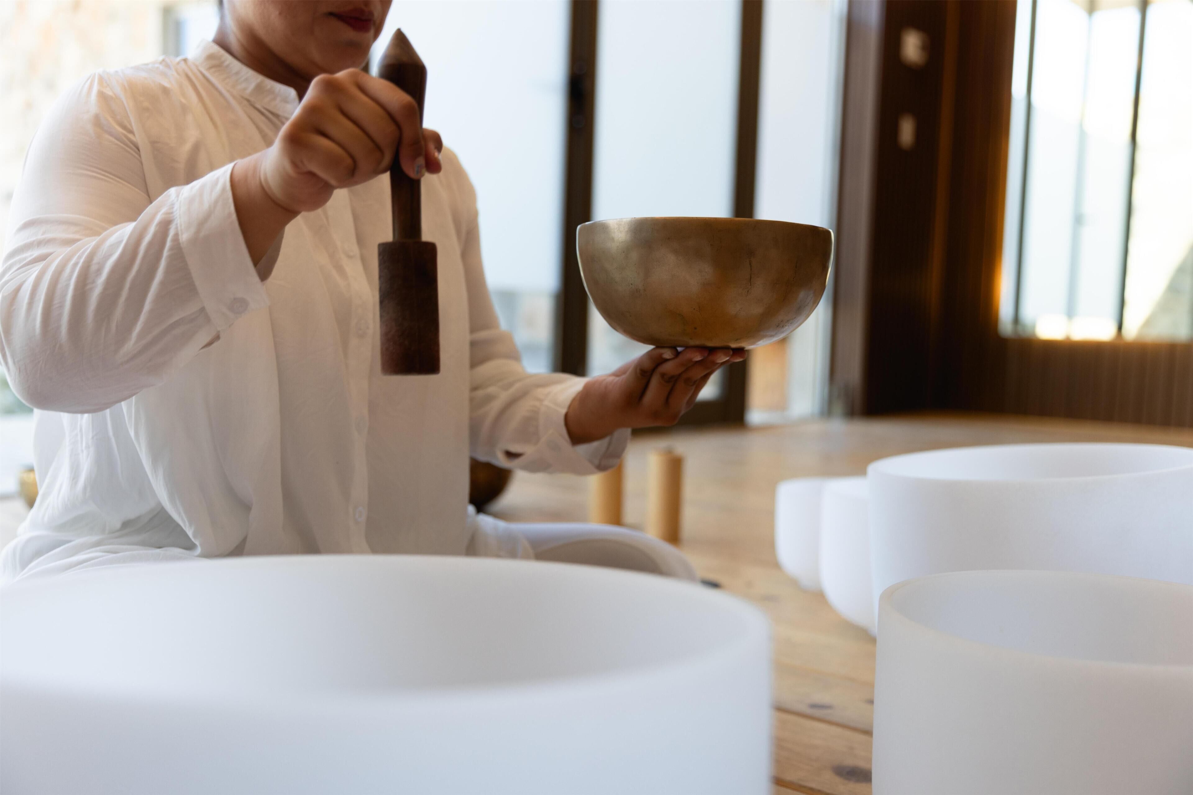 A person sitting on a white cushion, holding a wooden mallet and a large bowl, likely for meditation or gong practice, in a room with wooden flooring and white cushions.