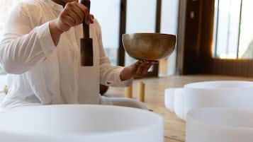 A person sitting on a white cushion, holding a wooden mallet and a large bowl, likely for meditation or gong practice, in a room with wooden flooring and white cushions.