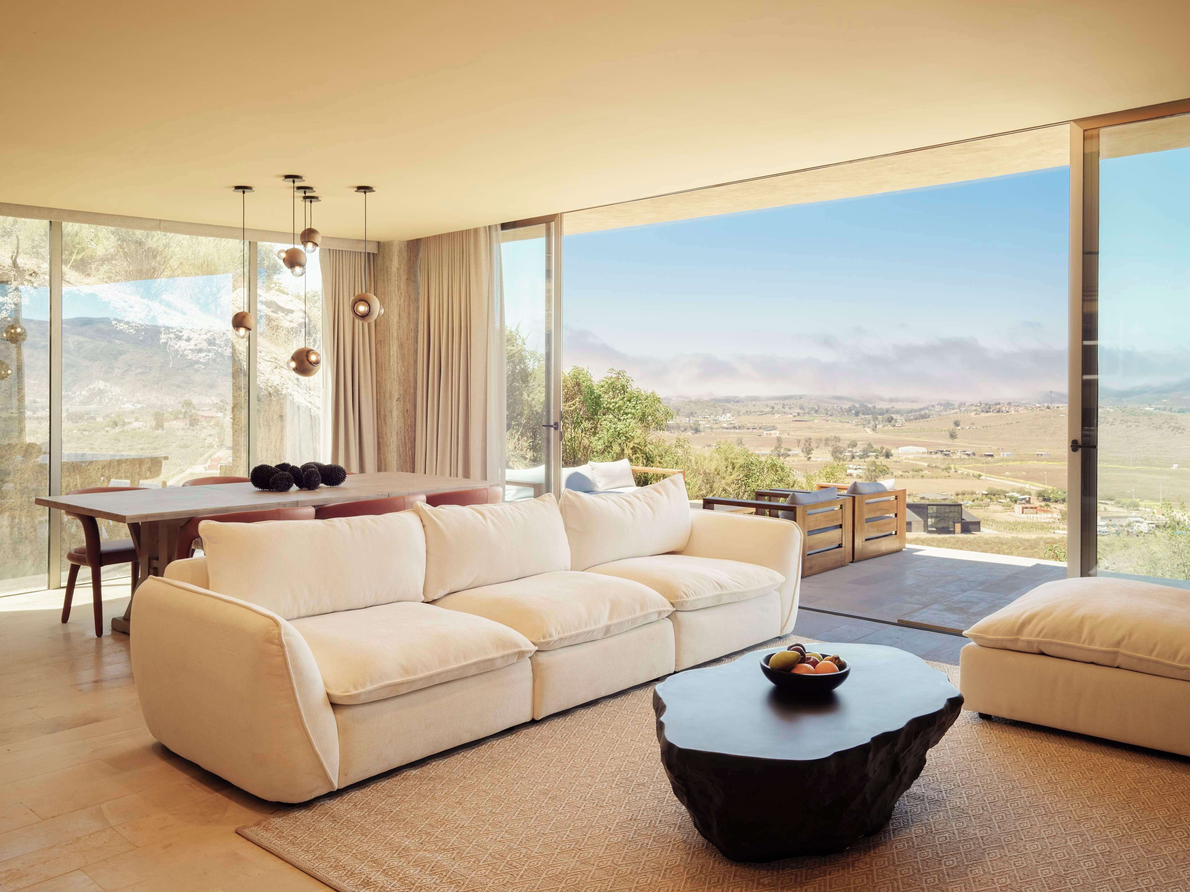 A spacious living room with a large sofa, a dining table, and a coffee table. There is a view of a mountainous landscape through large windows.