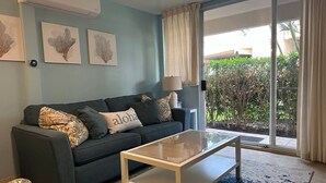 Una sala de estar con un sofá azul oscuro, una mesa de centro de cristal y un tapete estampado. Hay tres obras de arte colgadas en la pared y una ventana con cortinas.