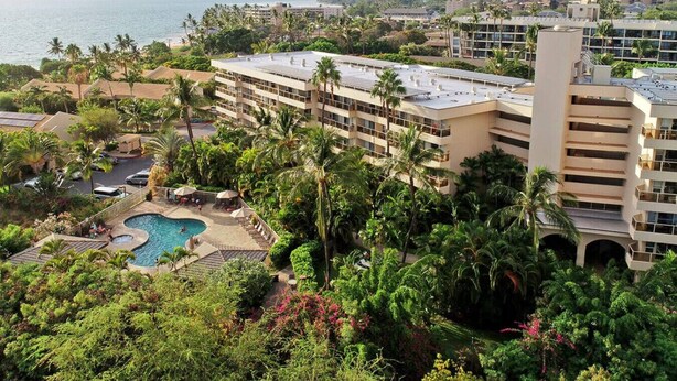 Un hotel de varios pisos, piscina y abundante vegetación.