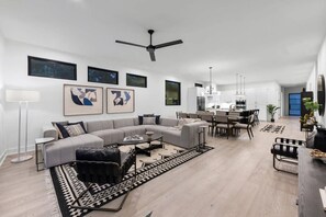 Un'ampia zona giorno con un divano modulare grigio, un tavolino da caffè e un tappeto decorato. C'è anche una sala da pranzo con tavolo e sedie, e una cucina sullo sfondo.
