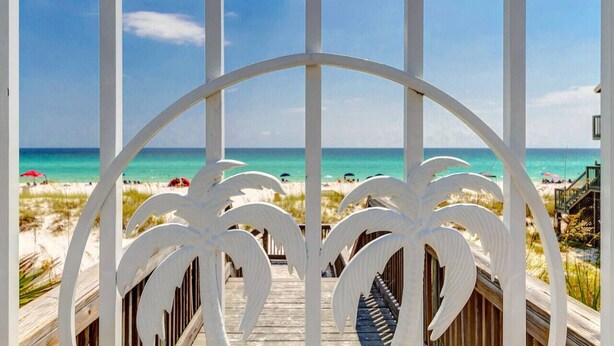 Una terrazza in legno con ringhiera bianca e motivo a foglia di palma, con vista su una spiaggia con persone e ombrelloni.