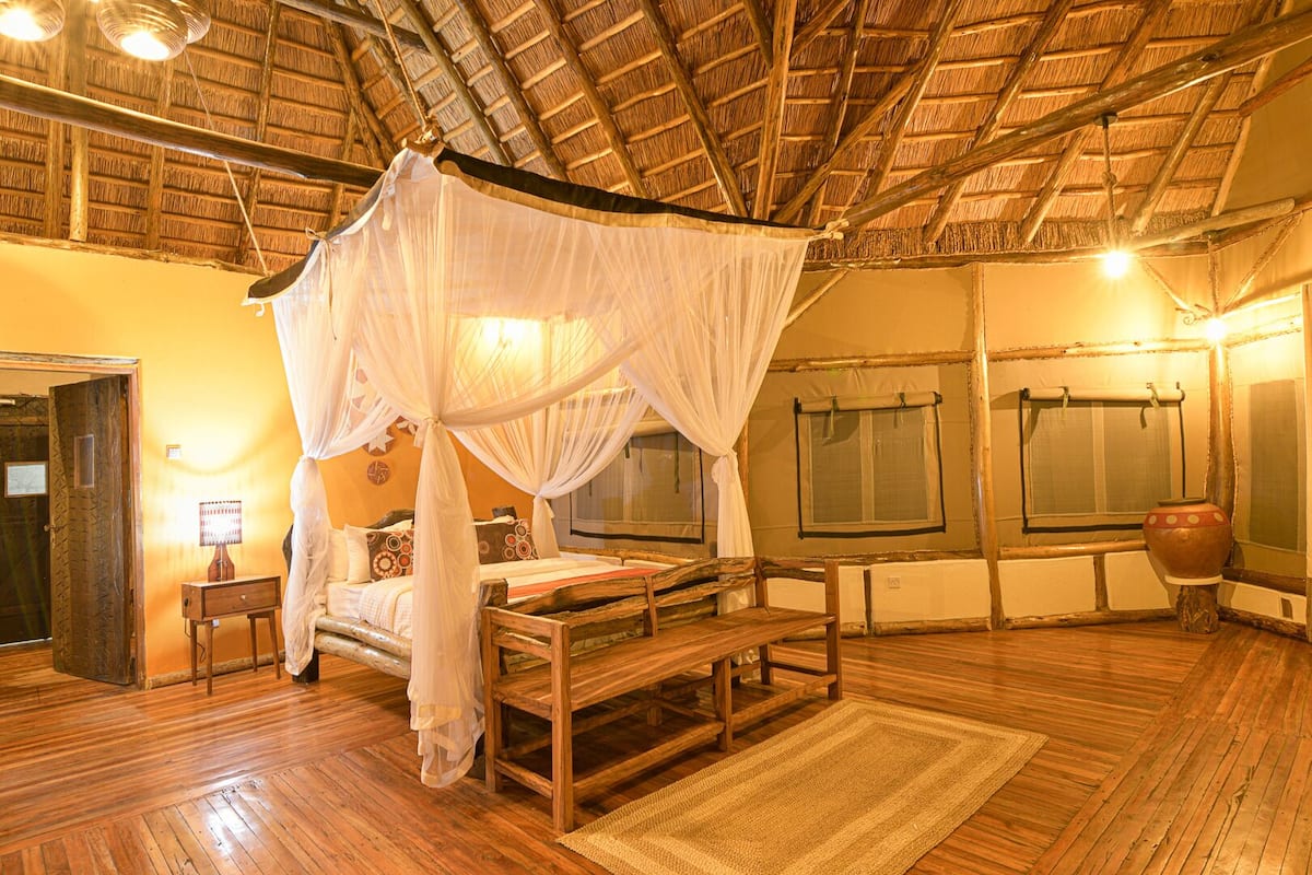 A room with a wooden bed under a canopy, wooden floors, and a thatched roof.