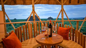 A wooden table with a bottle of wine and a glass, set against a backdrop of a thatched-roof structure and a scenic view. - Aardvark Safari Lodge (Kasese)