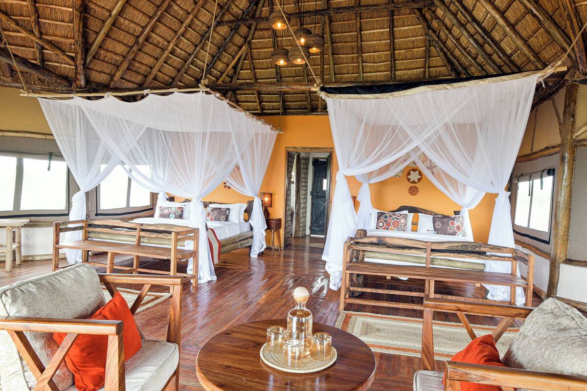 A room with two beds, each covered with white mosquito nets, wooden furniture, and a round table with a tray of glasses.