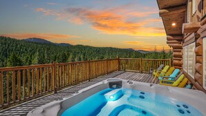 Um deck de madeira com banheira de hidromassagem e espreguiçadeiras, com vista para uma cordilheira arborizada ao pôr do sol.