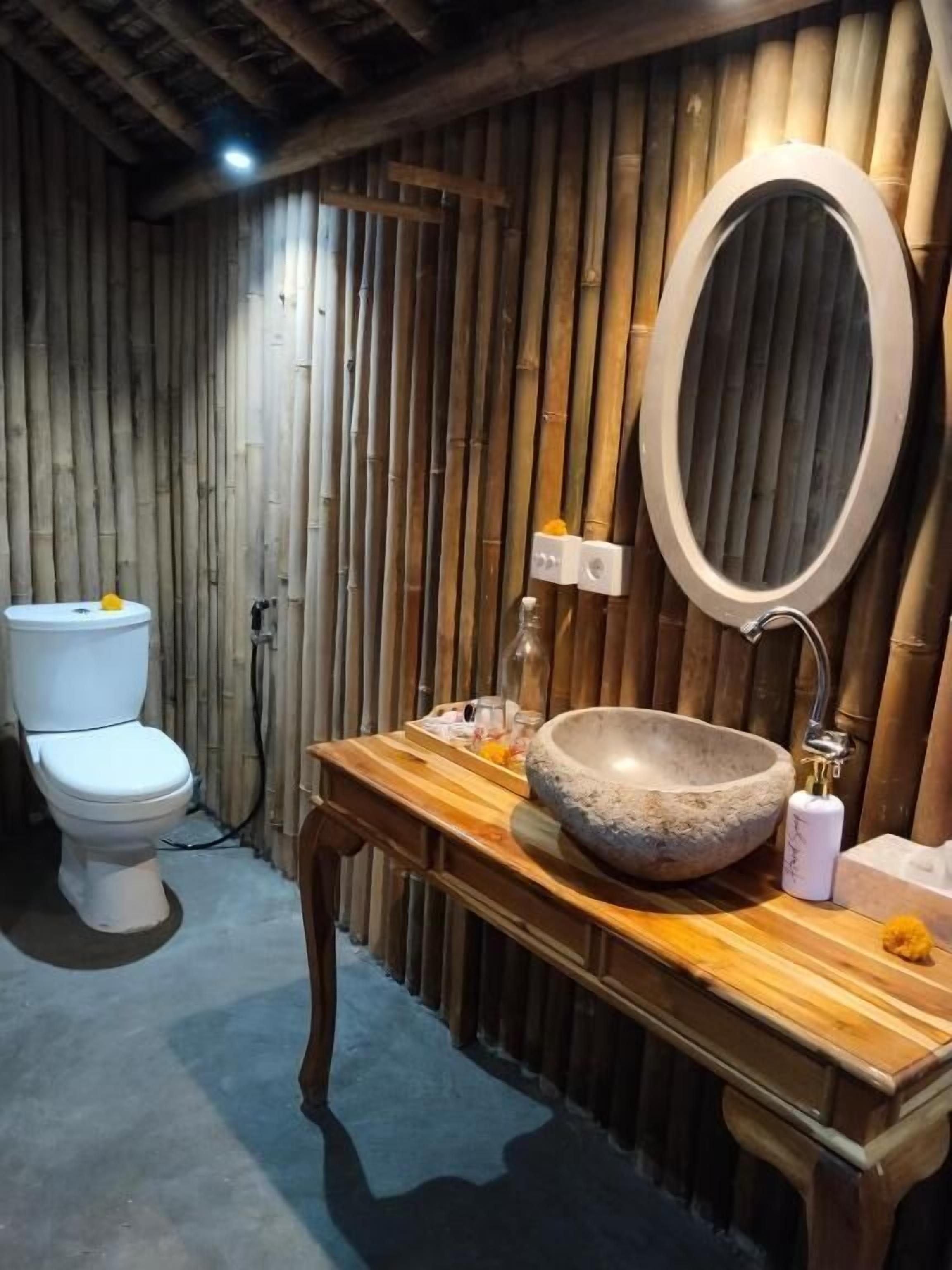A bathroom with a wooden vanity, a stone sink, a toilet, and a bamboo wall.