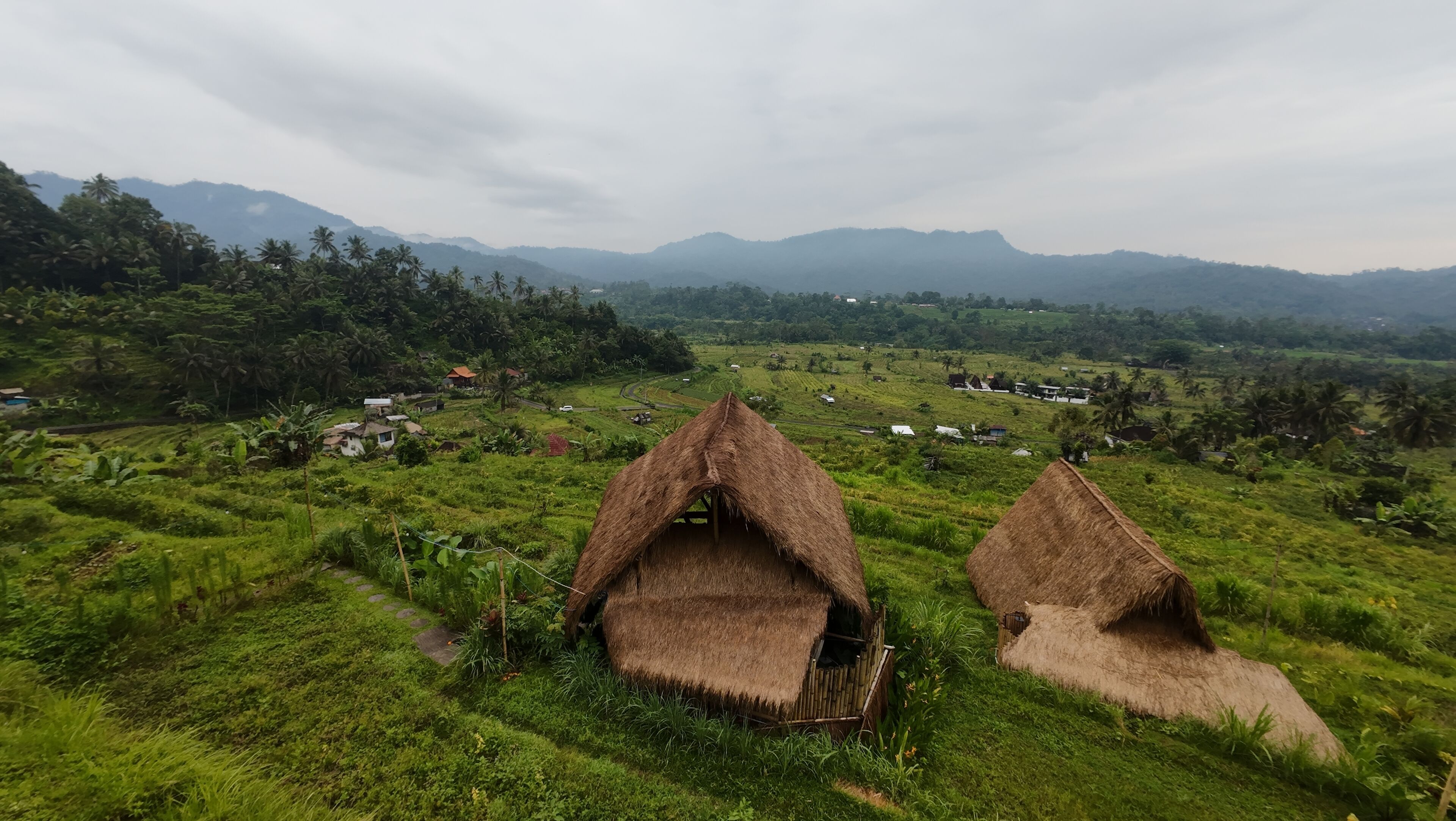 Two thatched-roof huts in a lush green valley with distant mountains.