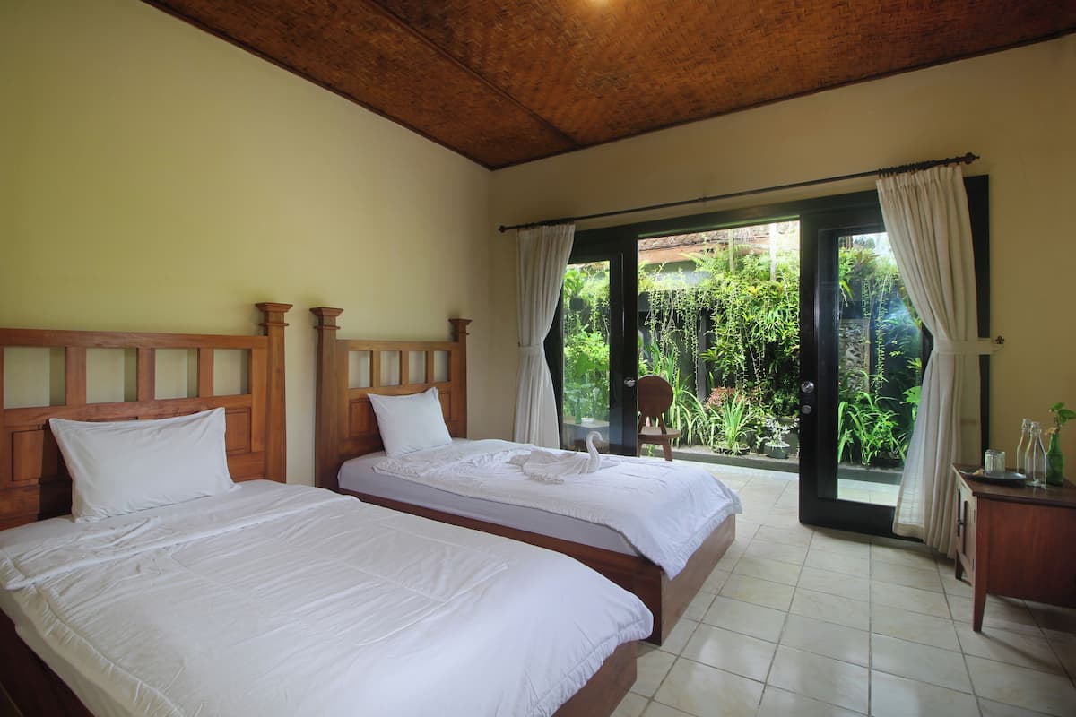 A bedroom with two beds, wooden headboards, and a view of greenery through a sliding glass door.