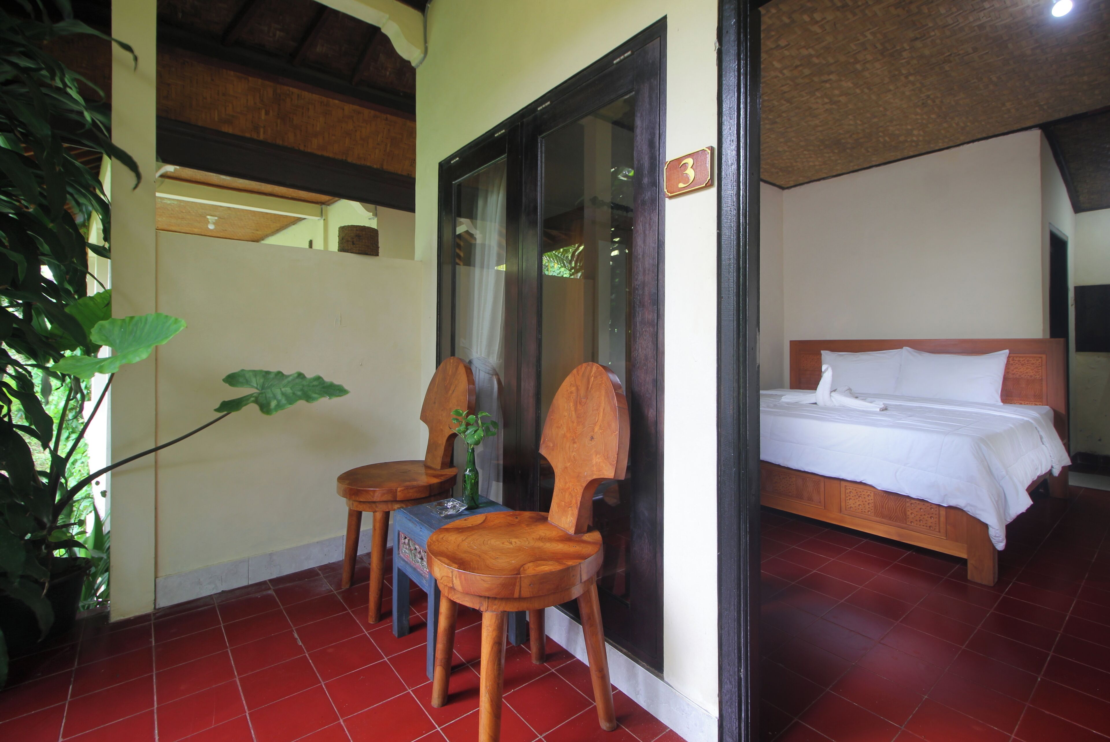 A room with a bed, two wooden chairs, a small table, and a potted plant.
