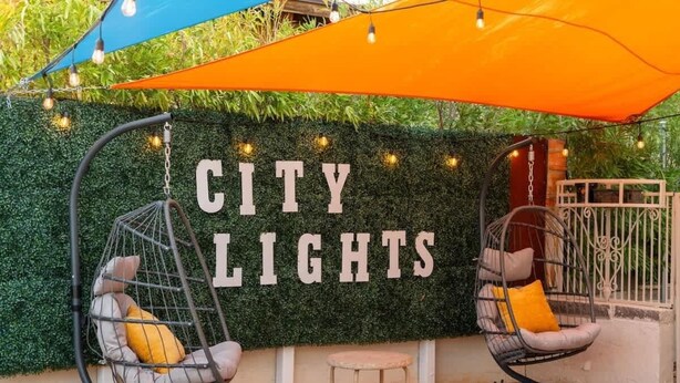Un área de descanso con dos sillones colgantes y una mesita, una pared con plantas y un letrero que dice 'CITY LIGHTS'.