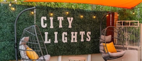 Un área de descanso con dos sillones colgantes y una mesita, una pared con plantas y un letrero que dice 'CITY LIGHTS'.