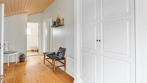 Un couloir doté d’un plancher en bois, de murs blancs et d’une grande porte blanche. On y trouve un banc avec un coussin et une petite étagère contenant des livres et des objets décoratifs.
