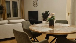 Un salon comprenant un canapé, une table de salle à manger ronde avec des chaises, un téléviseur et des plantes décoratives.