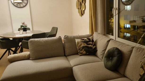 Un salon avec un canapé d’angle beige, une petite table ronde entourée de chaises et une fenêtre donnant sur des immeubles.