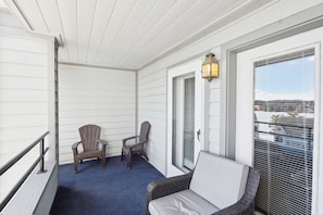 A covered balcony with two Adirondack chairs, a wicker chair, and a wall-mounted lantern.