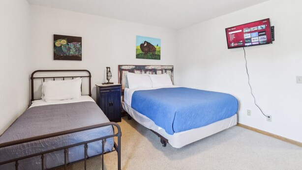 Un dormitorio con dos camas individuales, una mesita de noche, un televisor montado en la pared y dos cuadros colgados.