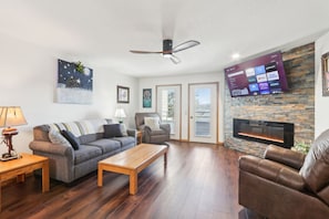 A living room with a fireplace, a flat-screen TV mounted on a stone wall, a ceiling fan, and a large window. - Wisconsin Dells Escape - Amazing Views - Pools (Wisconsin Dells)