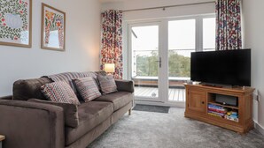 A living room with a brown sofa, patterned cushions, a television on a wooden stand, and a sliding glass door leading to a balcony. - River View (Preston)
