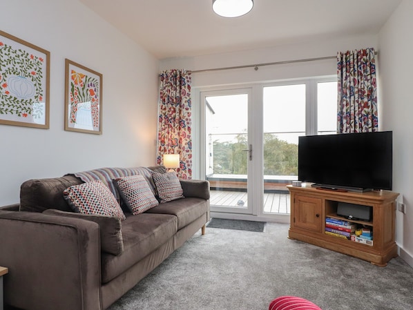 A living room with a brown sofa, patterned cushions, a television on a wooden stand, and a sliding glass door leading to a balcony.