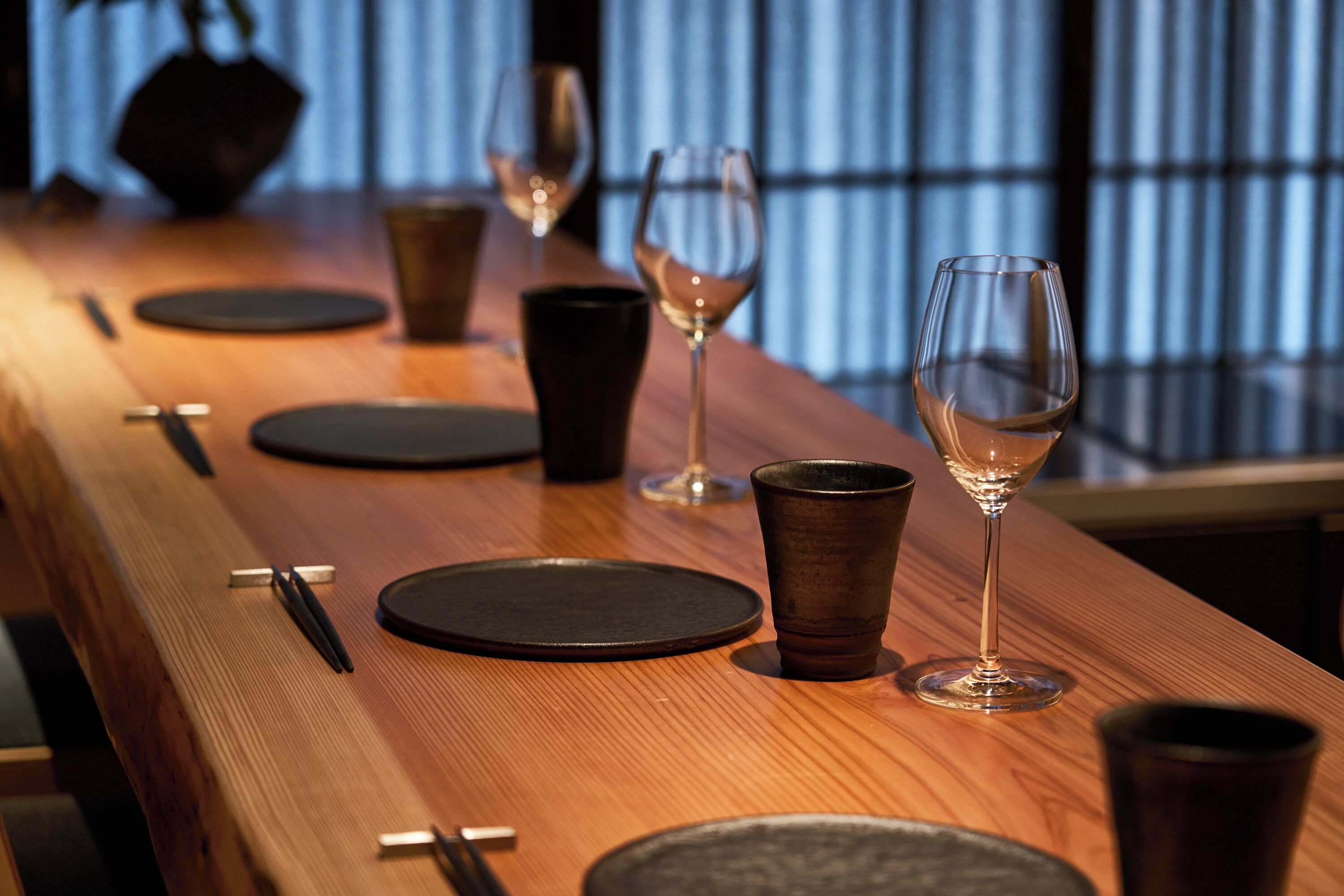 Una mesa de madera con platos negros, copas de vino y una taza oscura, frente a una ventana con persianas verticales.