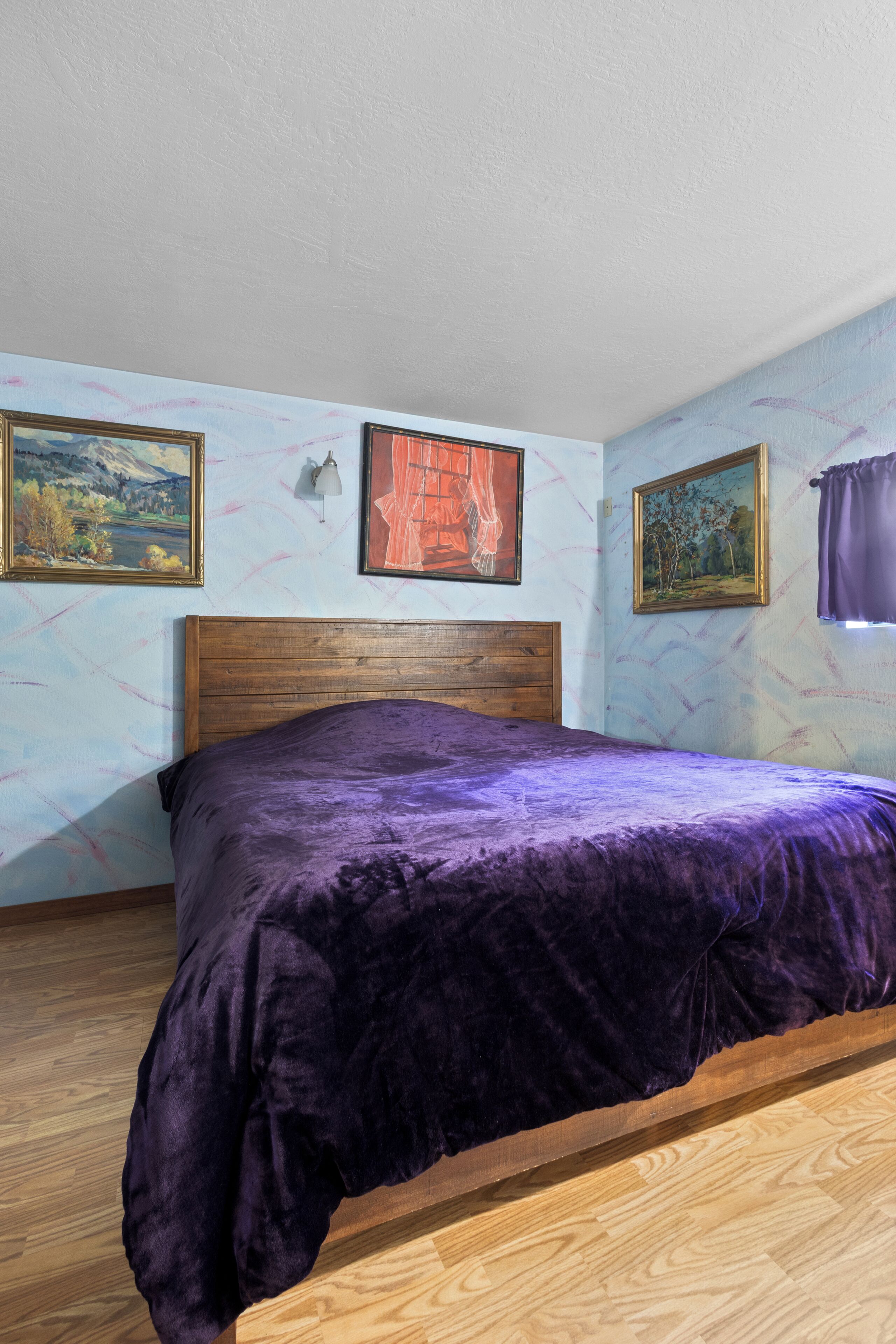 A bedroom with a wooden bed frame, a purple bedspread, and two framed paintings on the wall.