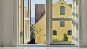 A view through an open window of a yellow building with a red roof and a small balcony, overlooking a body of water.