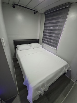 A single bed with a white mattress and pillows, a dark headboard, a window with blinds, and a light switch on the wall.