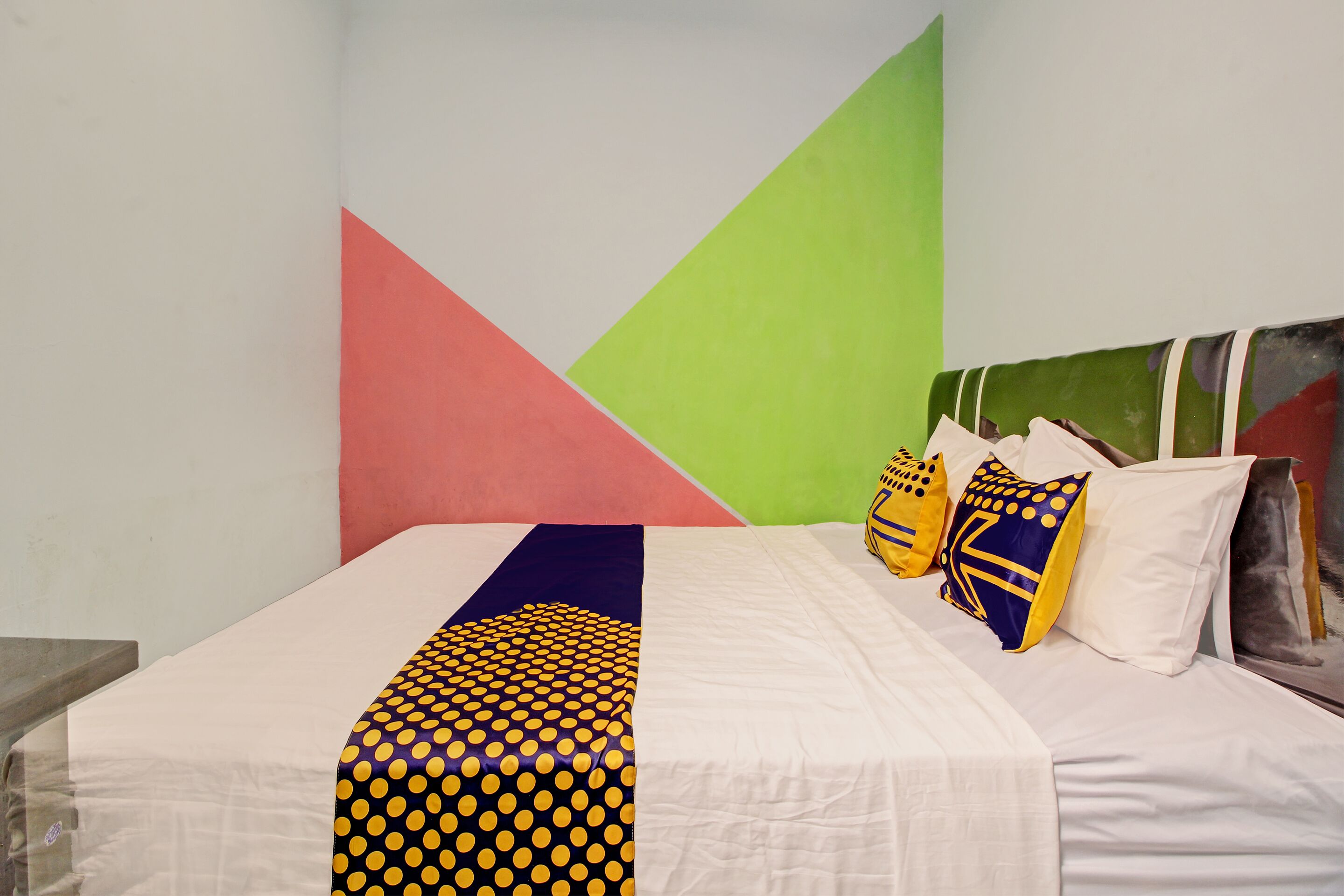 Una cama con sábana blanca y manta de estampado geométrico. Hay almohadas con diseños azules y amarillos. La habitación tiene una pared verde y rosa.