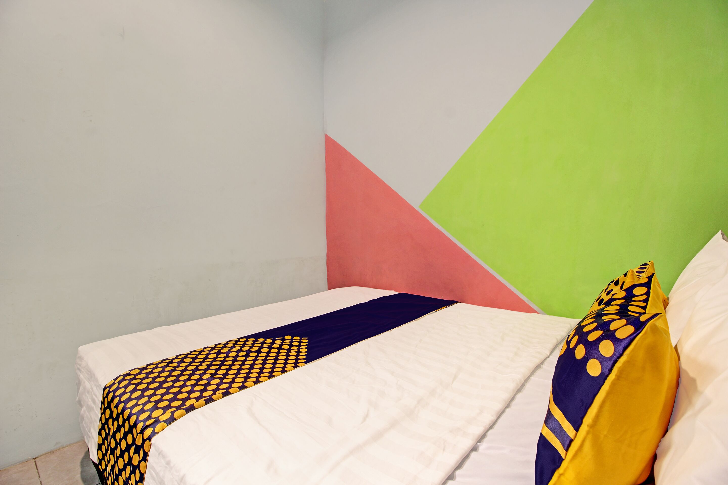 Een bed met een witte matras, een deken met een blauw-geel patroon en een kussen met een geel-blauw ontwerp. De kamer heeft een geometrisch wandontwerp met roze, groen en witte vlakken.