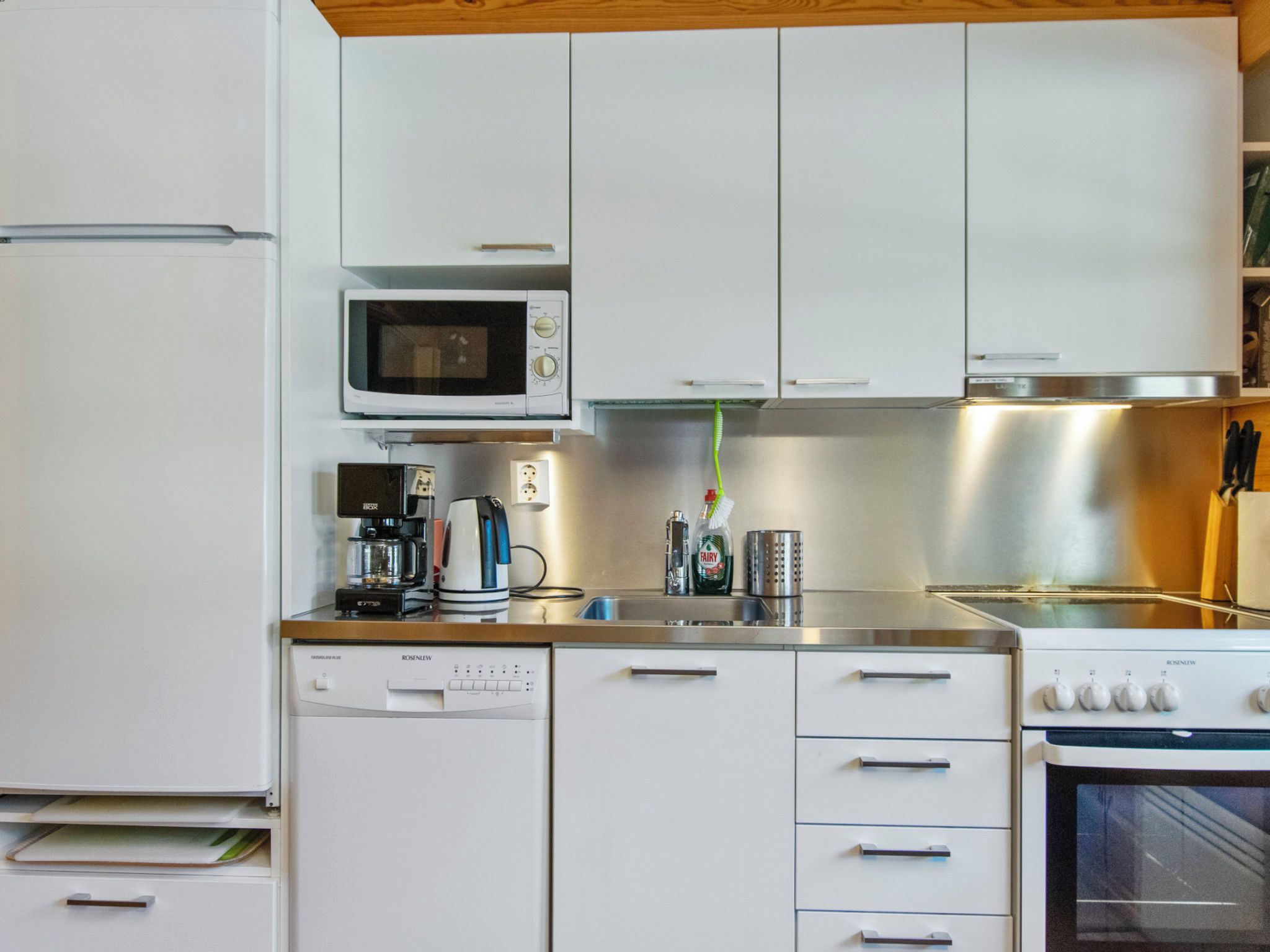 Una cucina compatta con mobili bianchi, un forno a microonde, una macchina per il caffè, un tostapane e un'lavastoviglie.