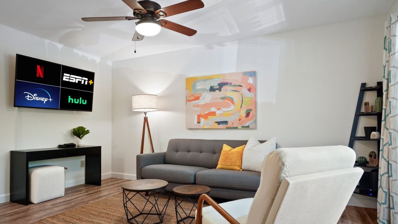 Un salon moderne avec un canapé gris, un fauteuil blanc, une table basse, une table d’appoint, un lampadaire et un téléviseur fixé au mur, qui affiche différents services de diffusion en continu.