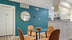 A dining area with a glass table and four chairs, a white door, a teal wall, a round mirror, and a kitchen with white cabinets and a microwave.