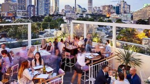 An outdoor dining area with tables and chairs, surrounded by greenery and overlooking a city skyline with various buildings and a tower. - Stylish Warehouse Oasis in Vibrant Darlinghurst (Darlinghurst)