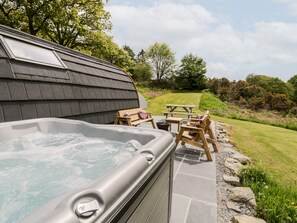A hot tub with a wooden bench and picnic table, surrounded by a stone patio and a grassy area with trees. - Llugwy Pod (Llanrwst)