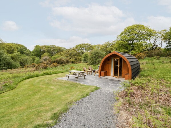 A cozy, semi-open wooden structure with a curved roof, a picnic table, and a bench, surrounded by a lush green landscape. - Llugwy Pod (Llanrwst)