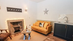 A cozy living room with a fireplace, a sofa, a coffee table, and a kitchen area with a sink. - Teal Cottage (Leyburn)