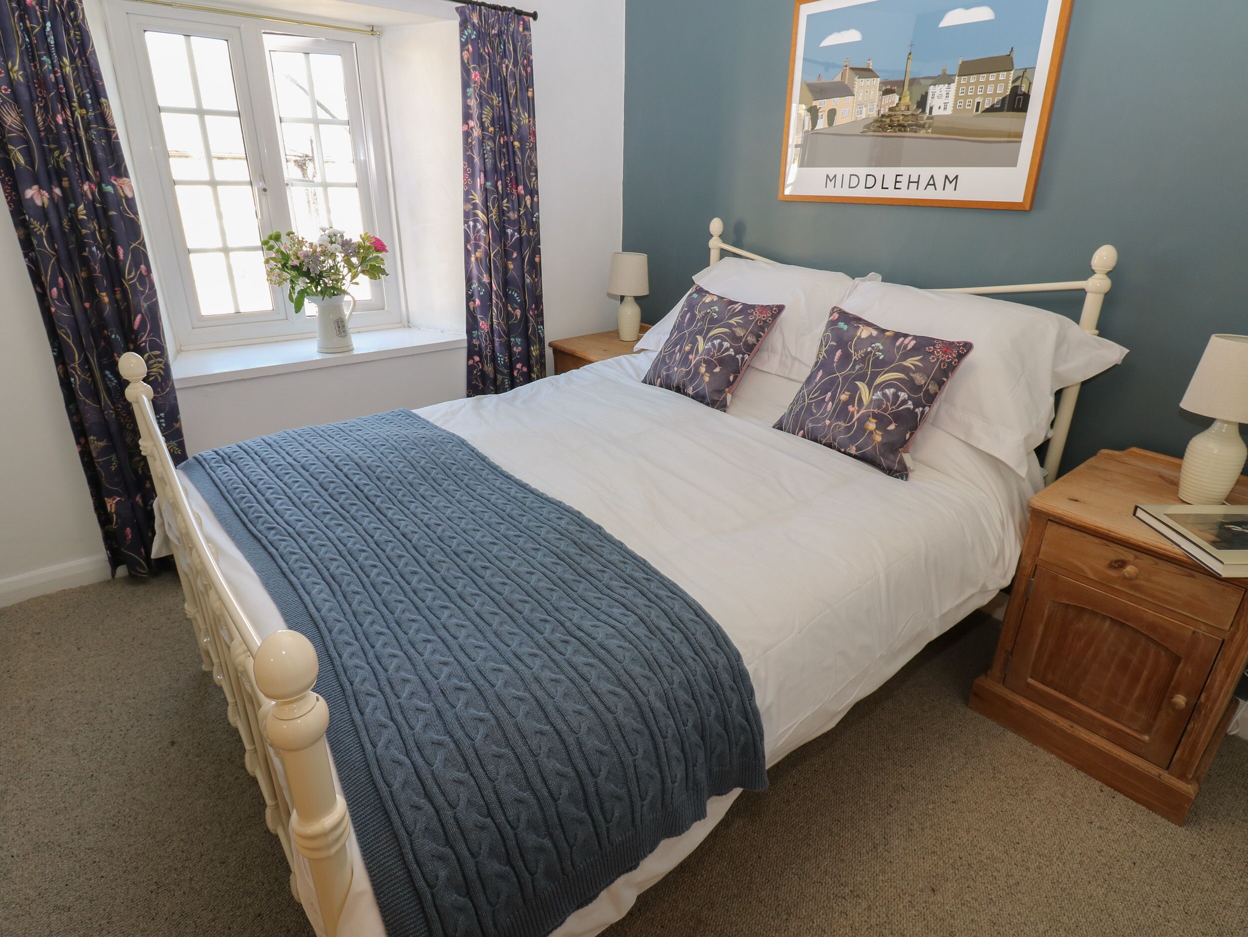 A neatly made bed with a blue blanket and floral pillows, a bedside table with a lamp, a window with curtains, and a framed picture of Middleham on the wall.