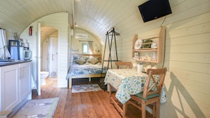 A cozy room with a bed, a table with a tablecloth, a chair, a television mounted on the wall, and a shelf with dishes.