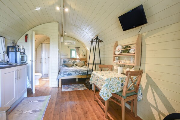 A cozy room with a bed, a table with a tablecloth, a chair, a television mounted on the wall, and a shelf with dishes. - Pendragon Pod (Axminster)
