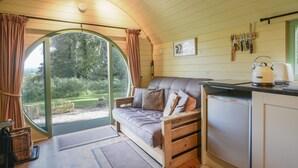 A cozy room with a curved ceiling, a sofa with cushions, a small kitchen area with a kettle, and a view of a garden through a large window.
