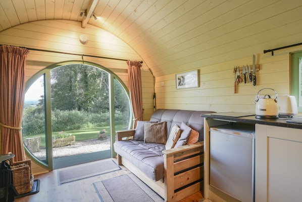 A cozy room with a curved ceiling, a sofa with cushions, a small kitchen area with a kettle, and a view of a garden through a large window. - Pendragon Pod (Axminster)