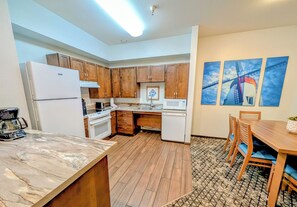 A modern kitchen with wooden cabinets, a refrigerator, stove, microwave, and a coffee maker. There is a dining area with a table and chairs.