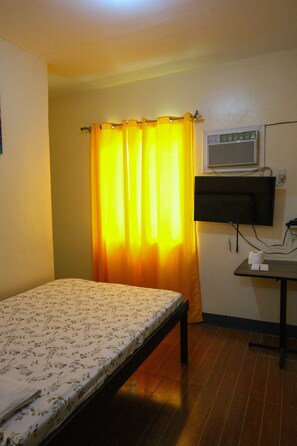 A small, single-bed room with a wooden floor, a bed with a floral bedspread, a television mounted on the wall, and a window with yellow curtains. - Villa Elsie Resort and Hotel (General Trias)